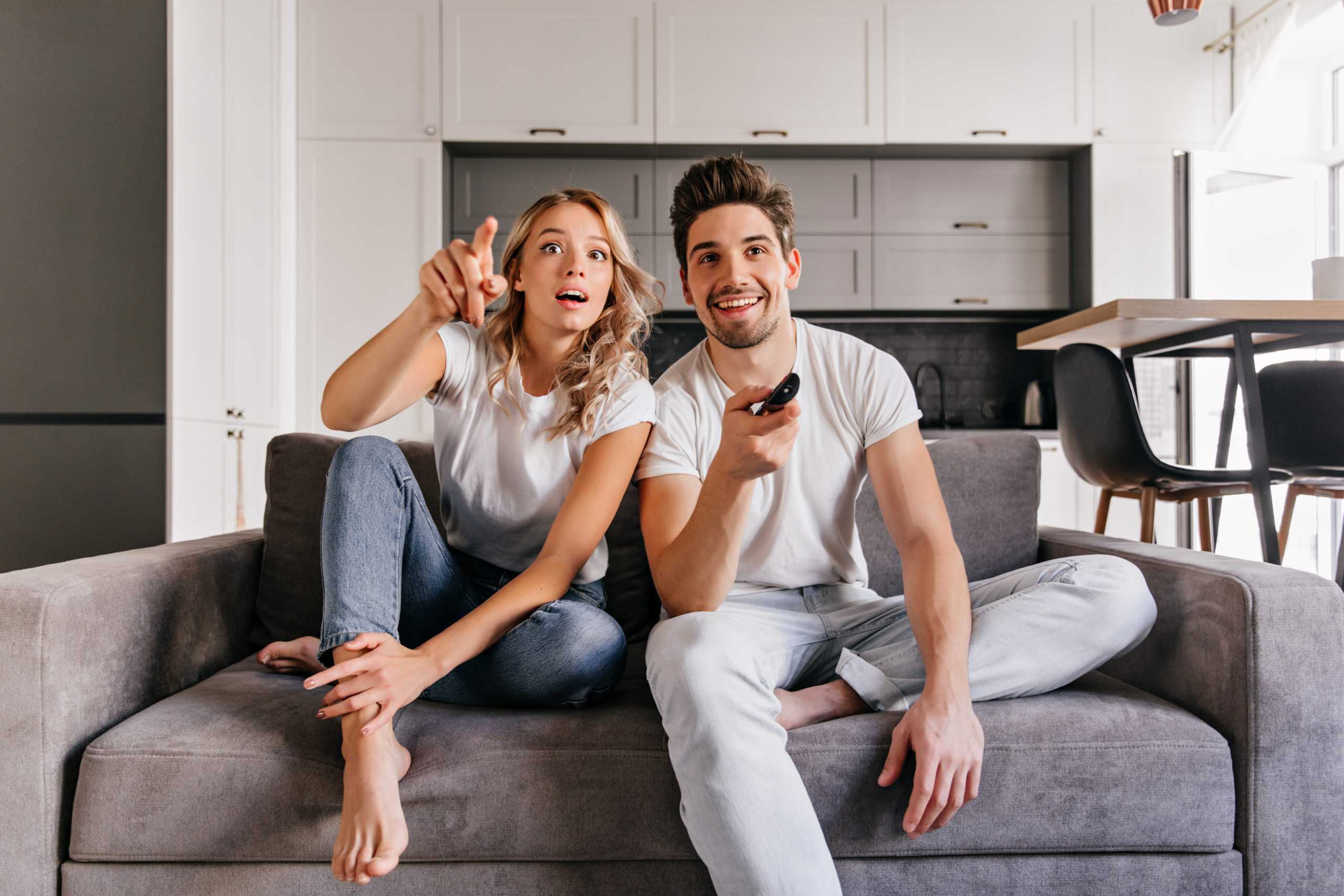 curious-couple-sitting-on-grey-sofa-indoor-portrait-of-man-and-woman-watch-tv-scaled