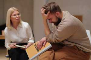 young-bearded-man-with-psychological-problem-or-trauma-holding-his-head-in-hand-while-sitting-by-mature-blond-female-counsellor-300x200